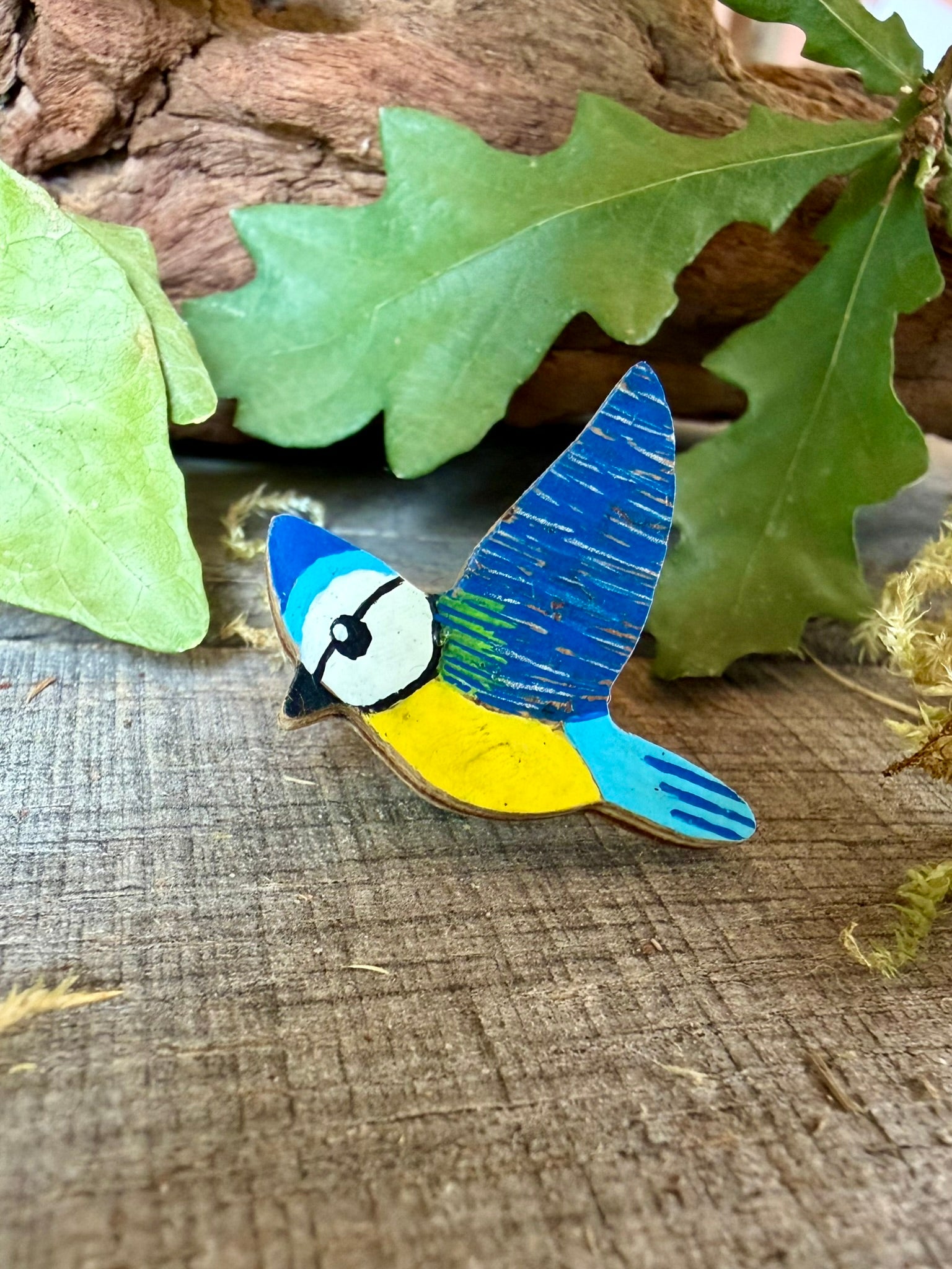 Handpainted blue tit cork pin badge on weathered wood with oak leaves and driftwood — sky blue cap, deep blue wings with silver line detail and yellow breast