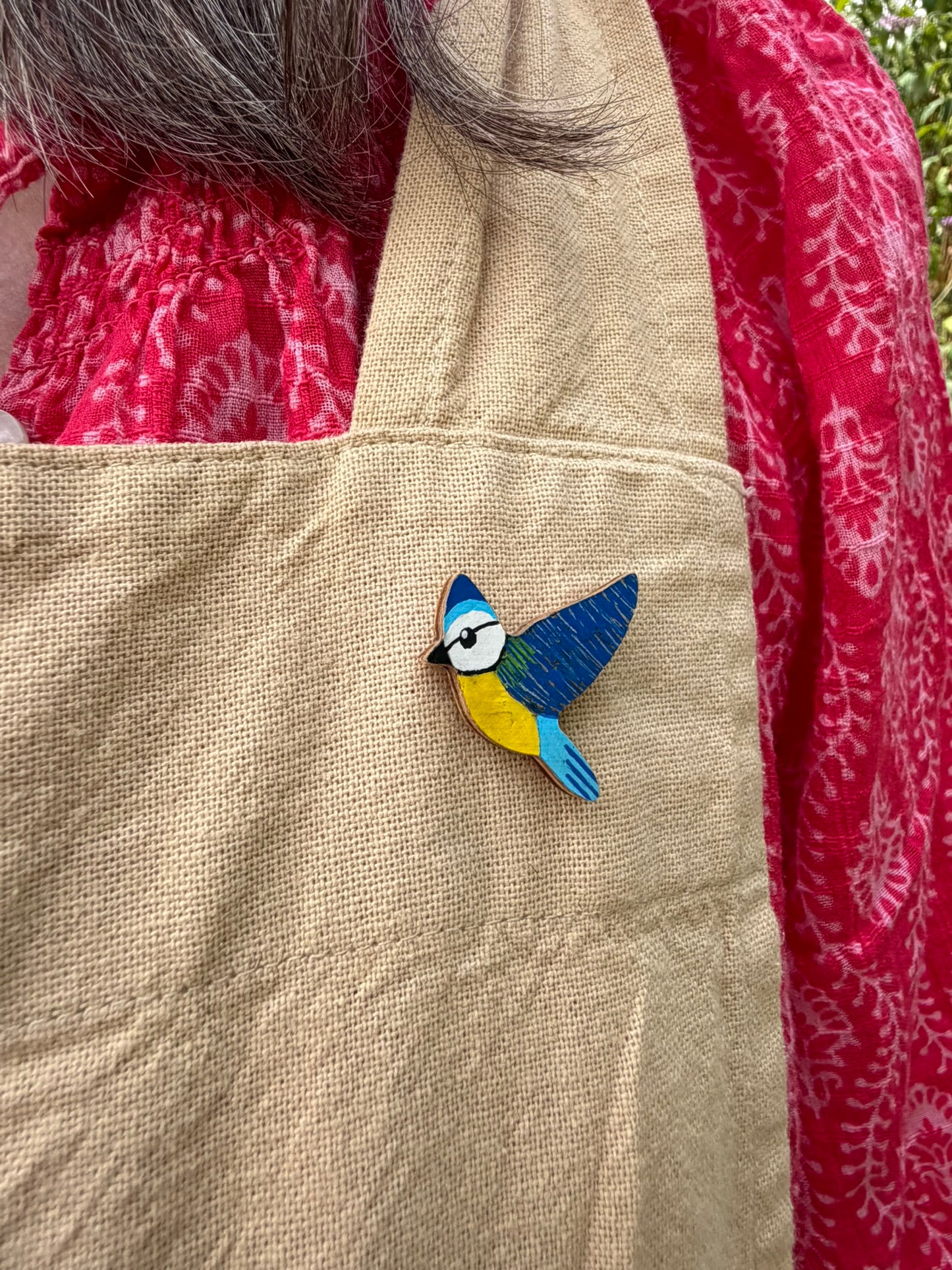 Blue tit cork pin badge pinned to a linen apron — handpainted sky blue and deep blue bird in flight, sustainable cork badge