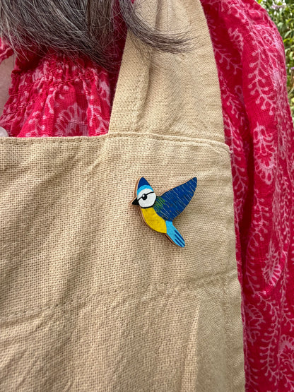 Blue tit cork pin badge pinned to a linen apron — handpainted sky blue and deep blue bird in flight, sustainable cork badge