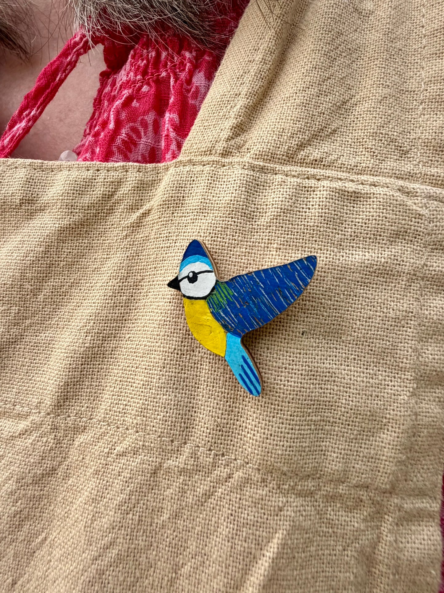 Blue tit cork pin badge pinned to a linen apron — handpainted sky blue cap, deep blue wings and yellow breast, sustainable cork badge handmade in Suffolk UK