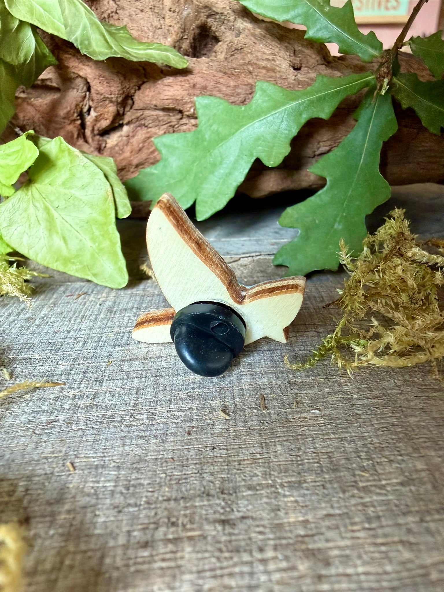 Reverse of the blue tit cork pin badge showing FSC-certified ply base and black rubber butterfly clasp — oak leaves and moss backdrop