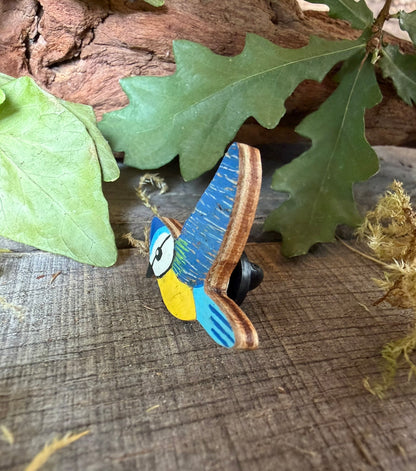 Side profile of the blue tit cork pin badge on weathered wood showing FSC ply base layers and black rubber butterfly clasp — oak leaves backdrop