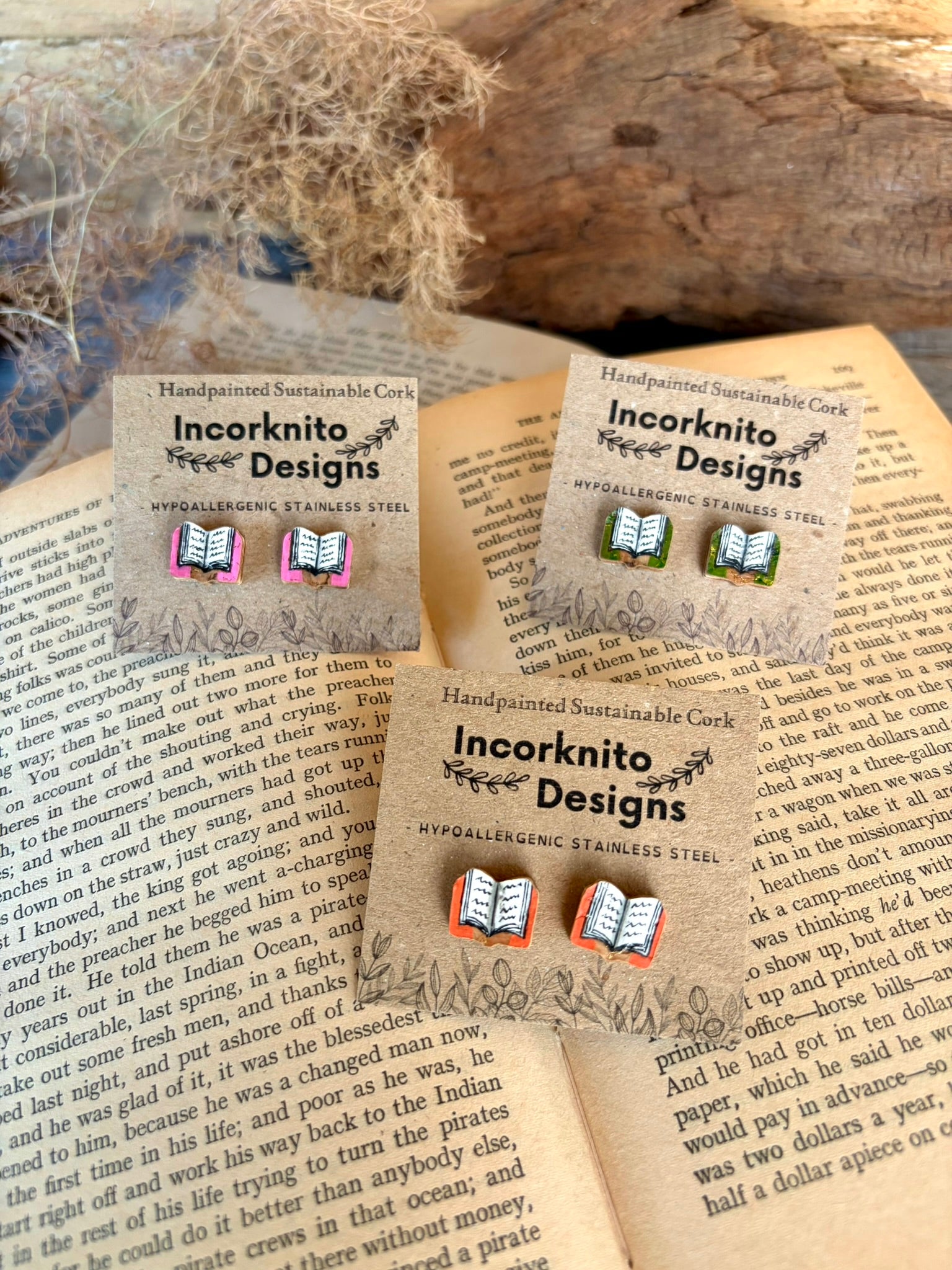 All three colour options of handpainted book cork stud earrings on an open book with driftwood and ivy — pink, orange and green