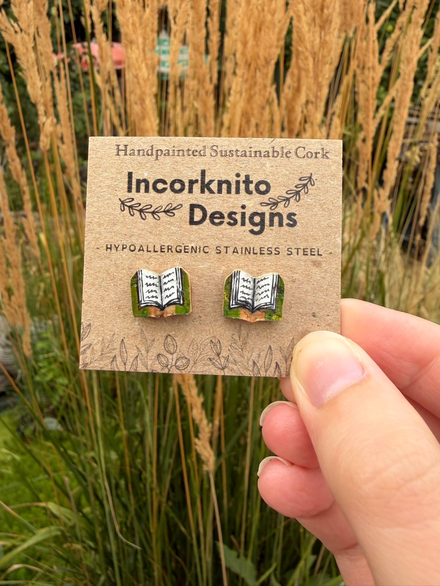 Green handpainted book cork stud earrings on an Incorknito Designs kraft card held against golden wheat grass