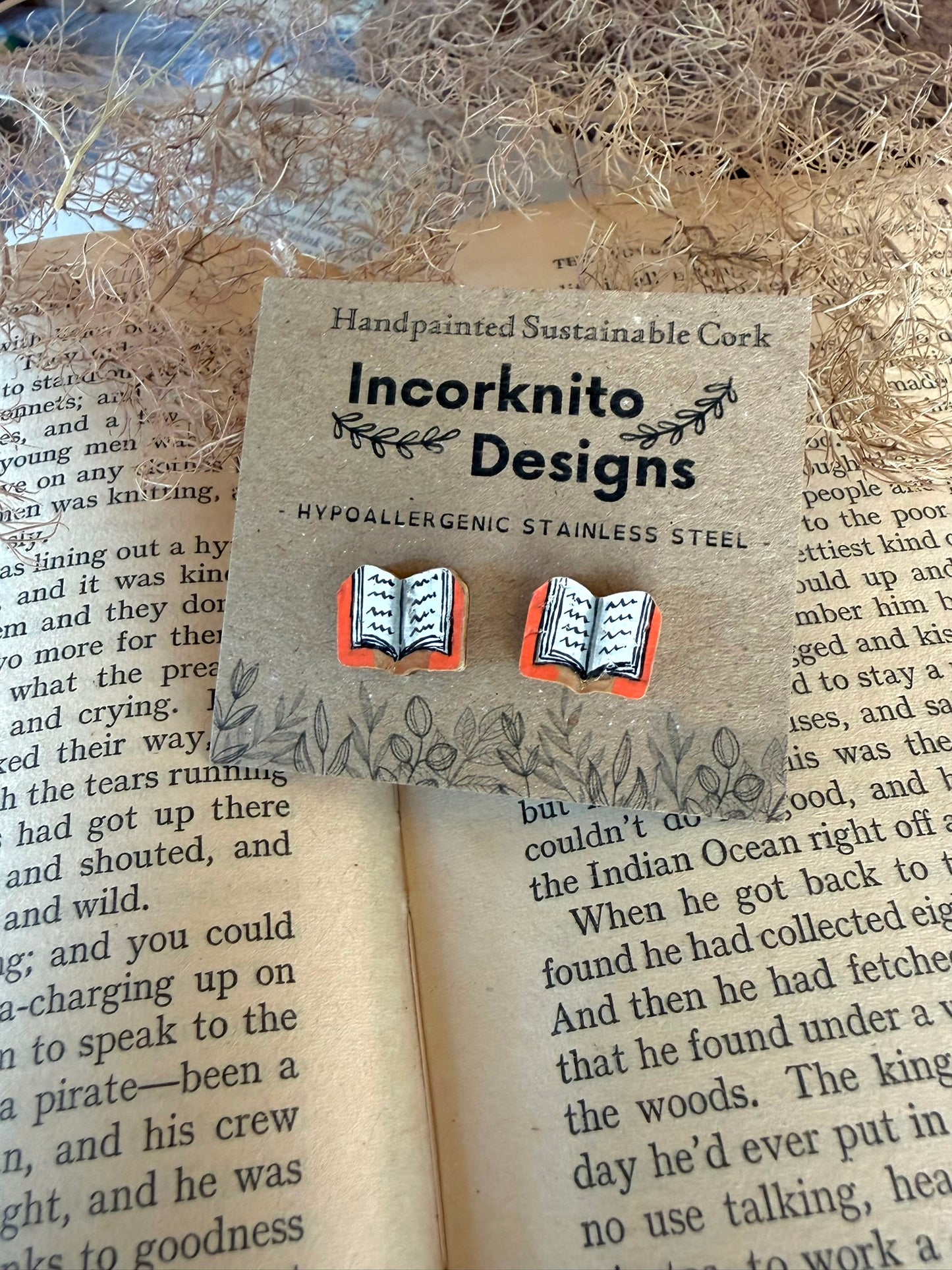 Orange handpainted book cork stud earrings on an Incorknito Designs kraft card laid on an open book with dried grass behind