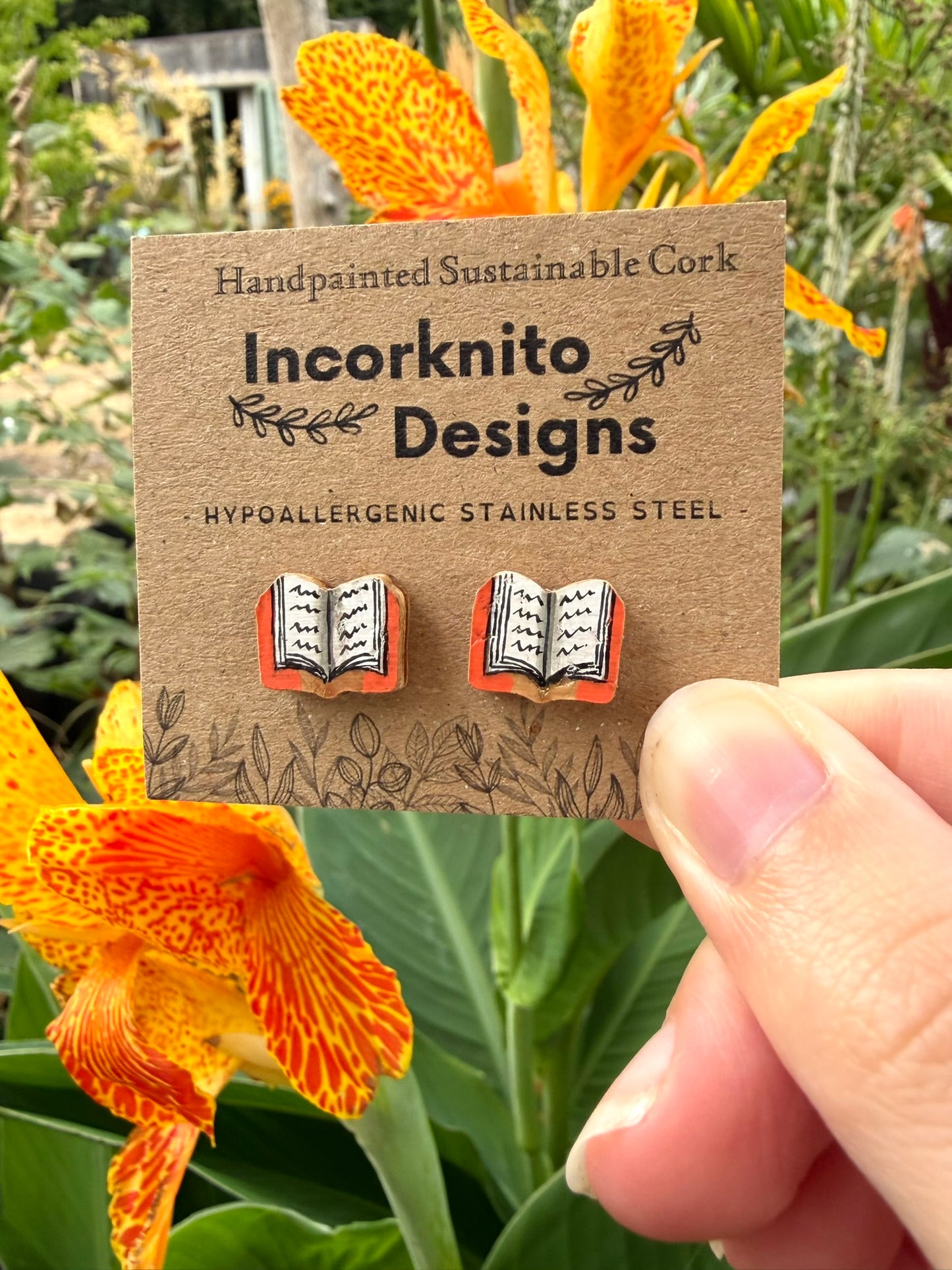 Orange handpainted book cork stud earrings on an Incorknito Designs kraft card held against vibrant orange canna lily flowers