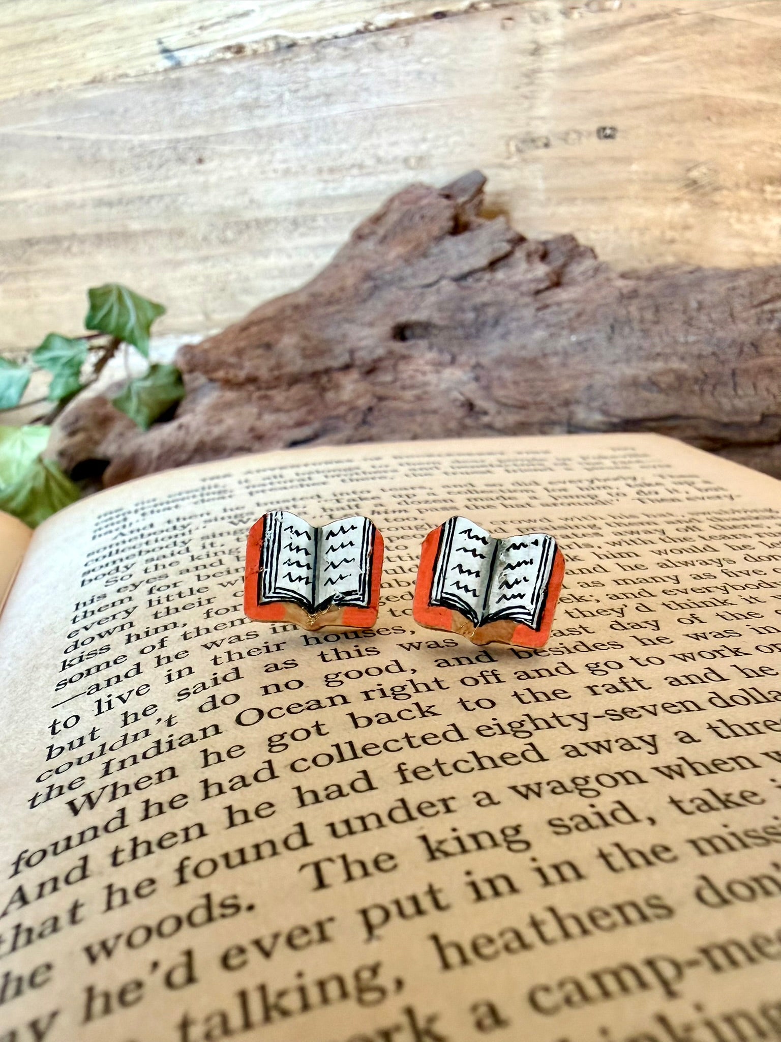Close-up pair of orange handpainted book cork stud earrings resting on the pages of an open book with driftwood and ivy behind