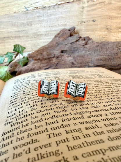 Close-up pair of orange handpainted book cork stud earrings resting on the pages of an open book with driftwood and ivy behind