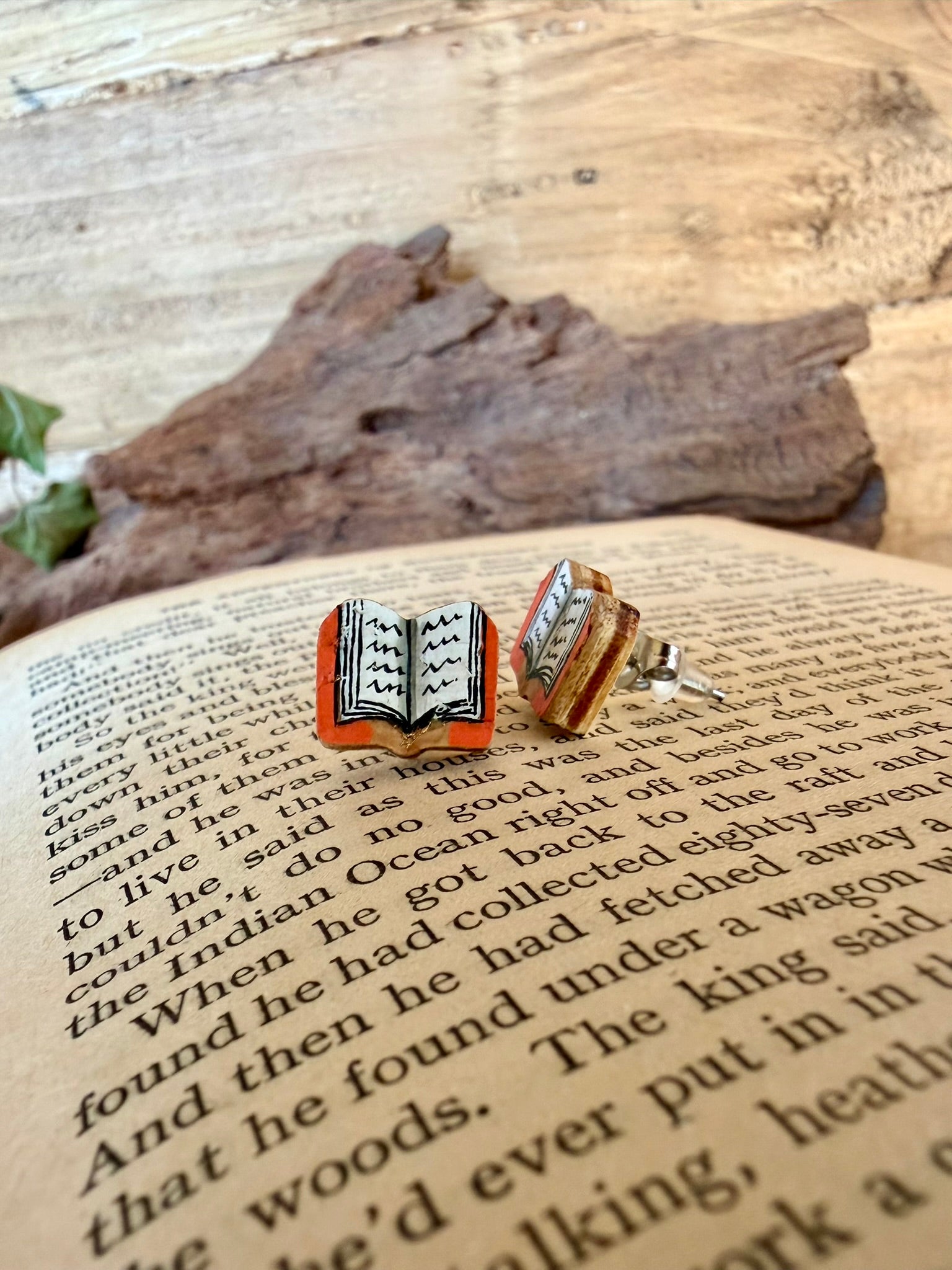 Side view of orange handpainted book cork stud earrings on an open book showing the natural wood reverse and stainless steel post, with driftwood and ivy behind