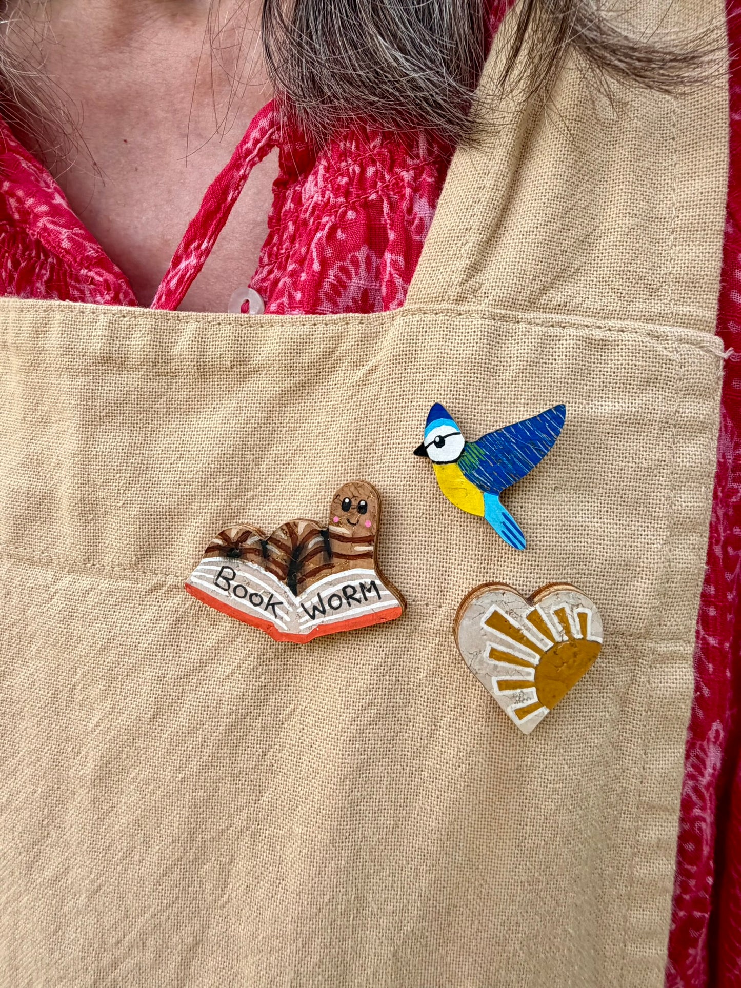 Natural bookworm, blue tit, and sun heart cork pin badges pinned to a linen apron — handpainted sustainable badges from Incorknito Designs