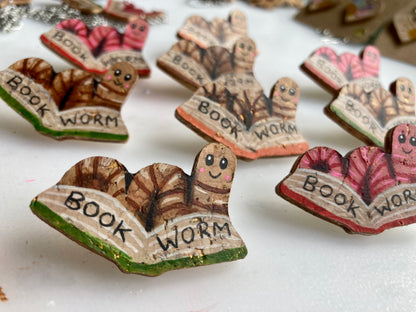 Flat lay of handpainted happy faced bookworm cork pin badges in pink and natural colourways on a workshop surface — also available in natural cork.