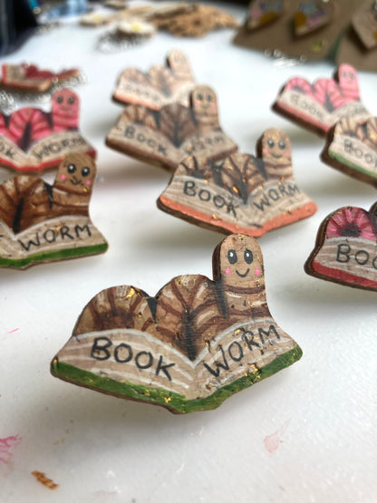 Multiple handpainted bookworm cork pin badges in progress on a workshop desk, showing both pink and natural cork colourways — handmade in Suffolk UK by Incorknito Designs