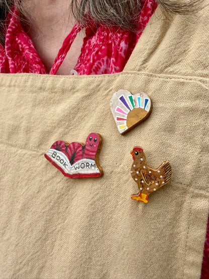 Handpainted pink bookworm, rainbow heart, and chicken cork pin badges pinned to a linen apron — handpainted sustainable badges from Incorknito Designs
