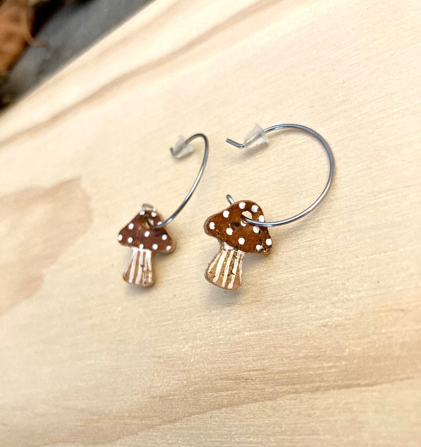 Brown toadstool cork mini hoop earrings hanging from a driftwood branch against a pale plywood wall — earthy tones with white spot detail