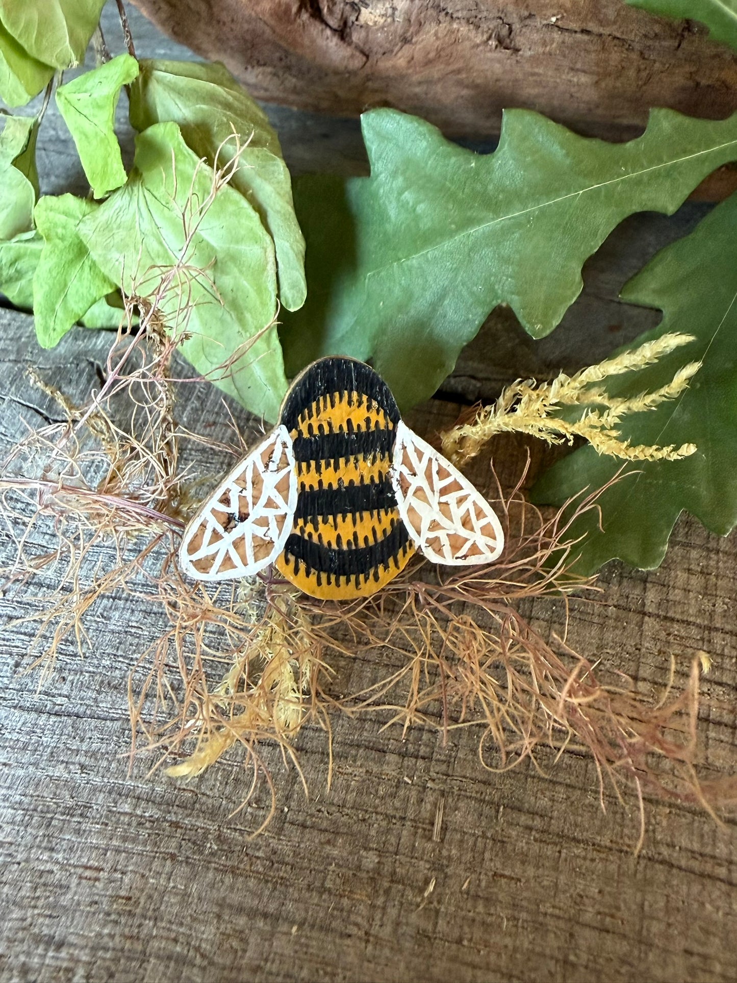 Handpainted bumblebee cork pin badge on weathered wood with dried grass, ivy and oak leaves — golden yellow and black stripes with white lace wings