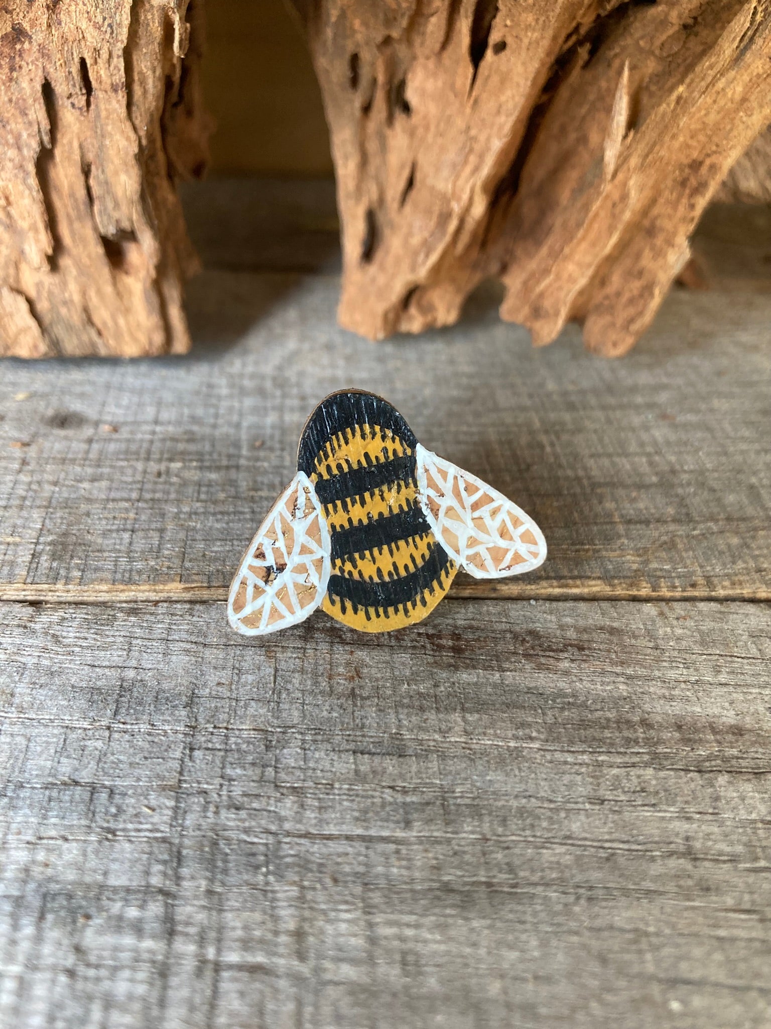 Handpainted bumblebee cork pin badge face-on on weathered wood with driftwood backdrop — golden yellow and black stripes with white lace wings, handmade in Suffolk UK