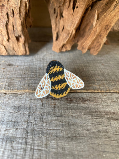 Handpainted bumblebee cork pin badge face-on on weathered wood with driftwood backdrop — golden yellow and black stripes with white lace wings, handmade in Suffolk UK