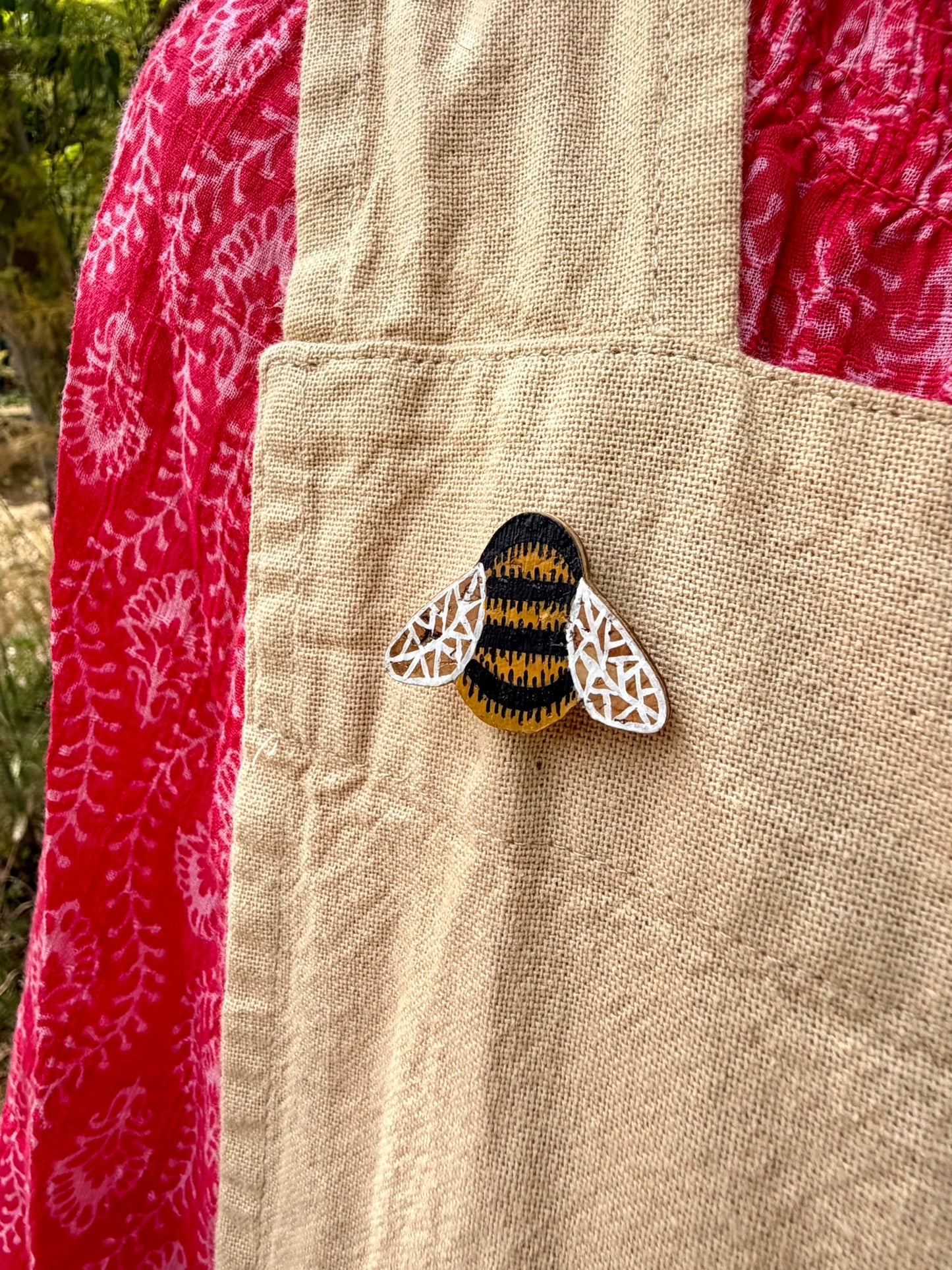 Bumblebee cork pin badge pinned to a linen apron pocket — handpainted golden yellow and black bumblebee with white lace wings, sustainable cork badge