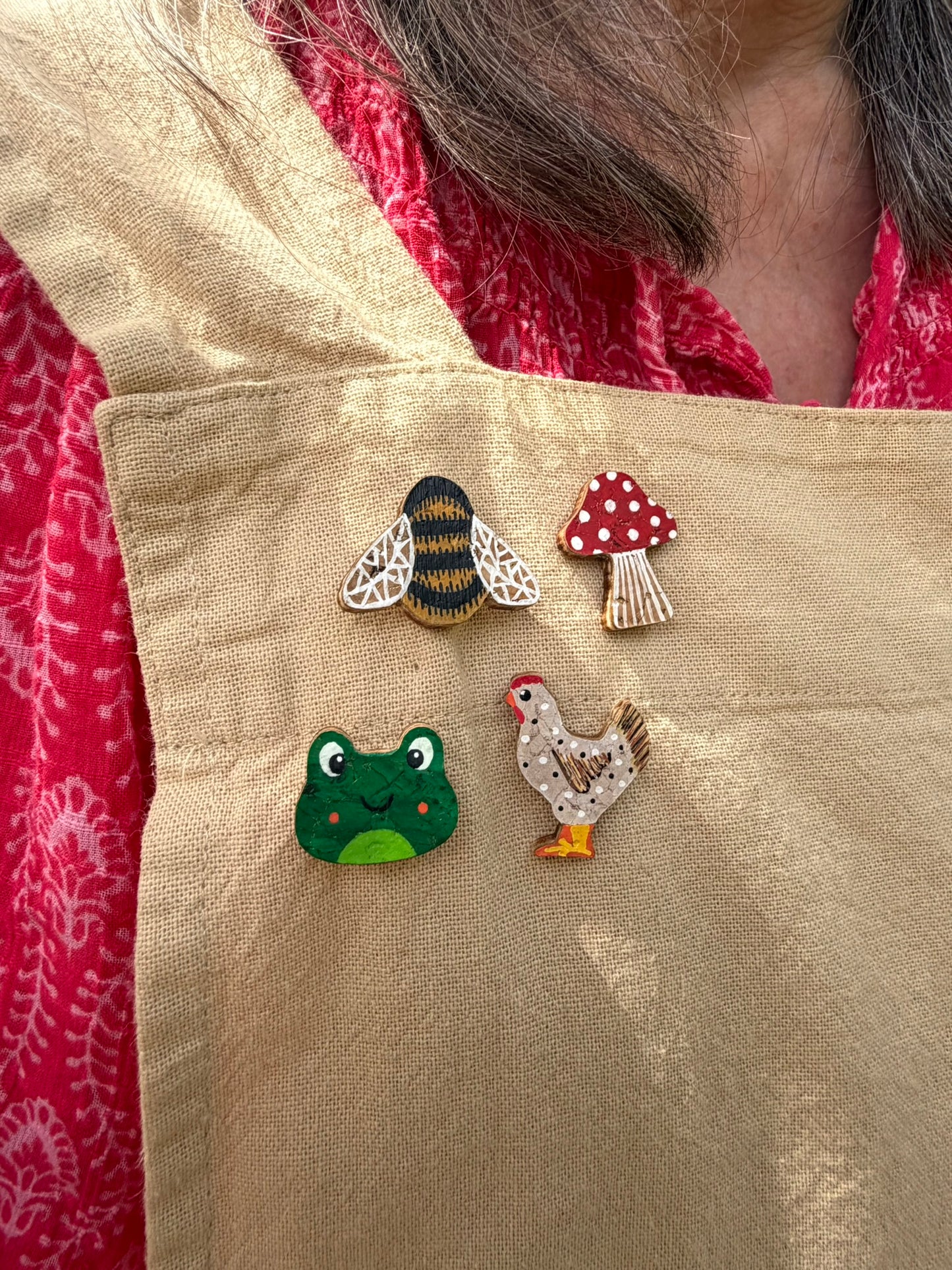 Bumblebee, red toadstool, frog, and chicken cork pin badges pinned to a linen apron — handpainted sustainable badges from Incorknito Designs