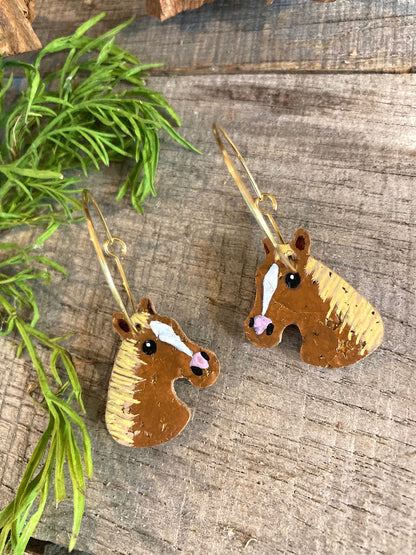 Chestnut tan horse cork earrings on gold hoops, flat lay on pale weathered wood with green foliage — eco-friendly handpainted horse jewellery Suffolk UK