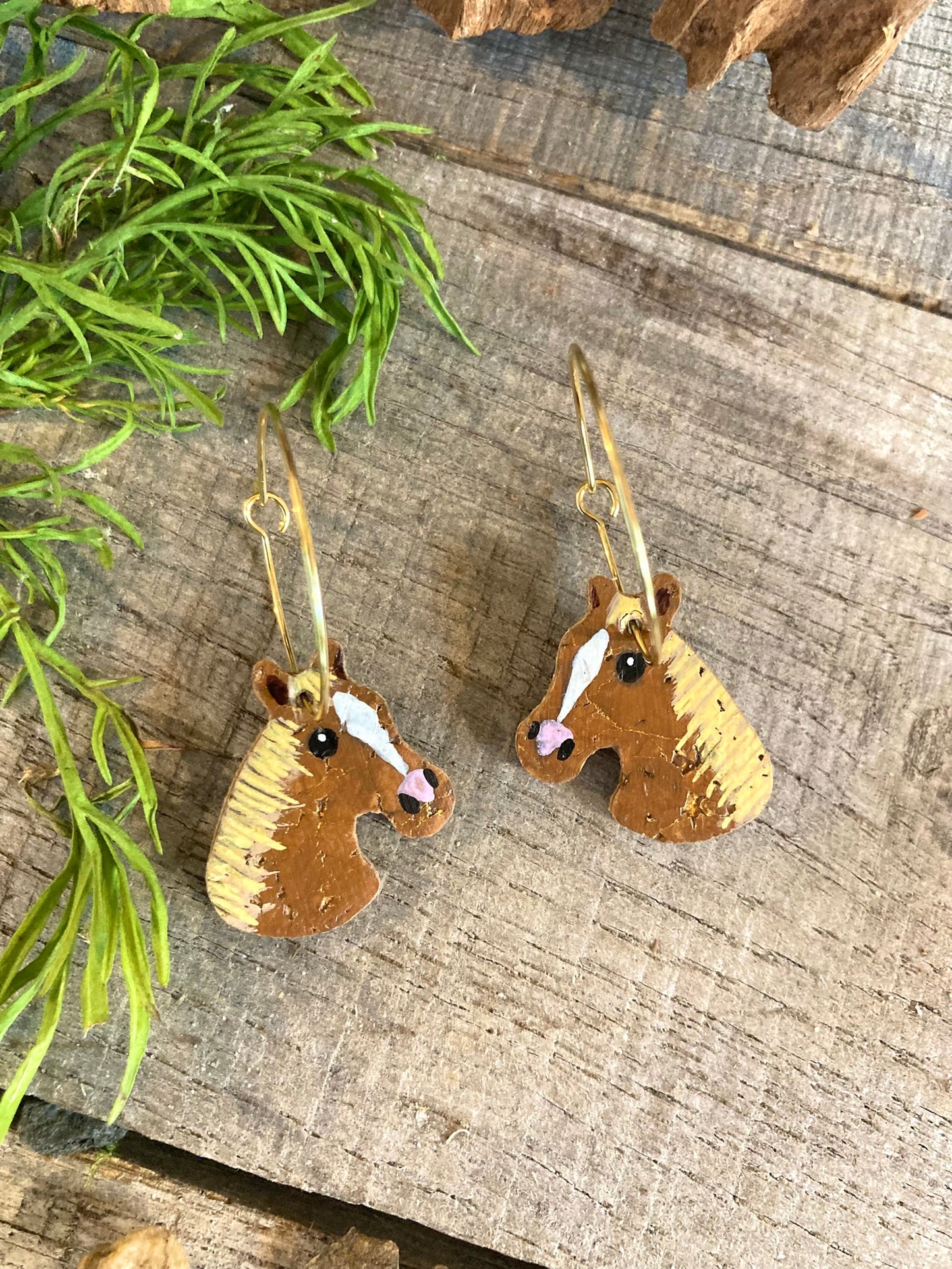 Chestnut and tan horse cork earrings on gold hoops, flat lay on weathered wood with green foliage — handpainted equestrian jewellery handmade in Suffolk UK