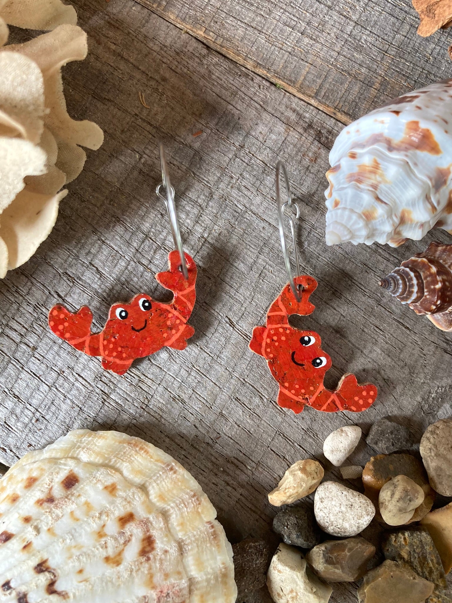 Cheerful red crab cork earrings on silver hoops, flat lay on weathered driftwood with seashells and pebbles