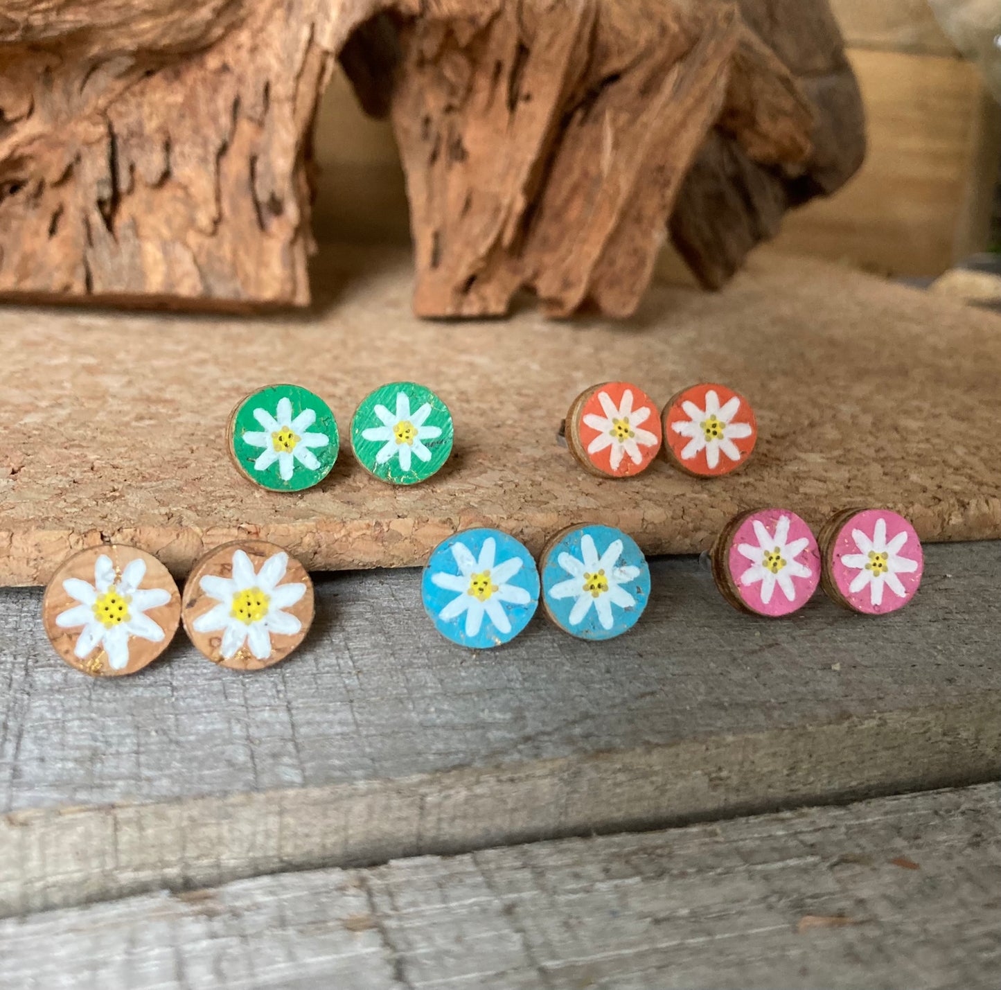 Handpainted daisy cork stud earrings in six colours — emerald, khaki, natural, orange, pink and light blue — on driftwood