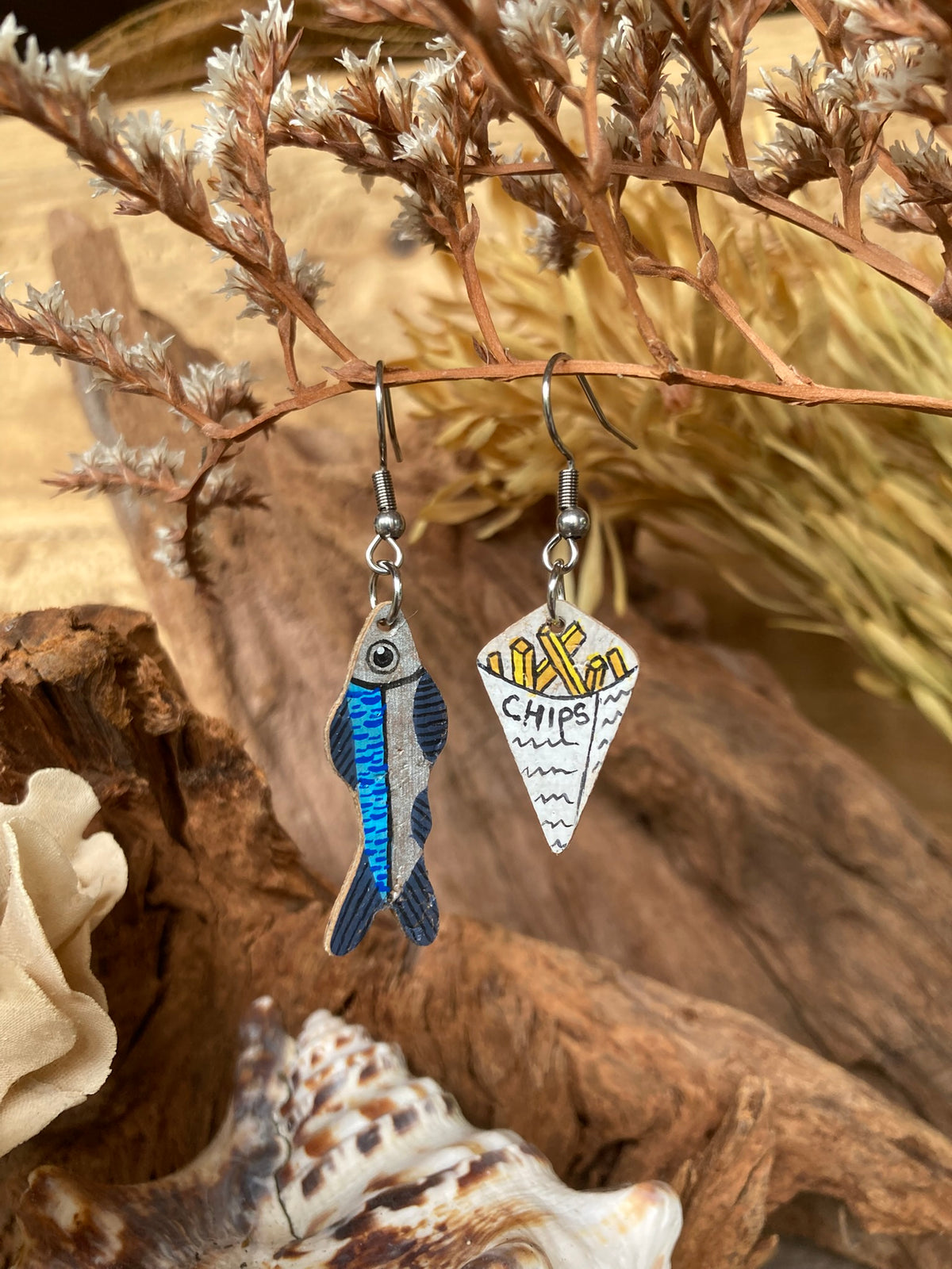 Handpainted cork fish and chips earrings hanging from dried botanicals — blue iridescent fish and white newspaper chip cone