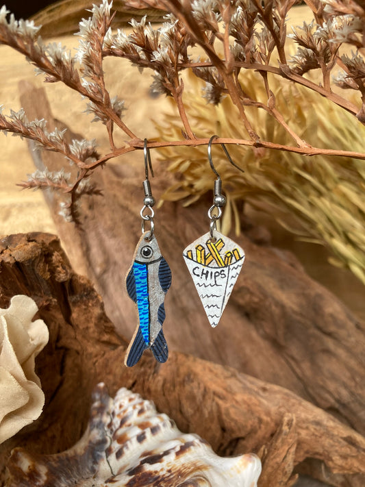 Handpainted cork fish and chips earrings hanging from dried botanicals — blue iridescent fish and white newspaper chip cone