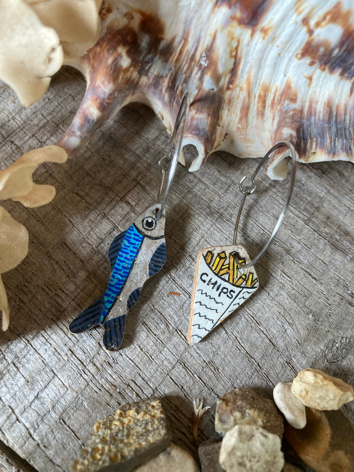 Cork fish and chips earrings on silver large hoops, laid on a driftwood surface with seashells and pebbles