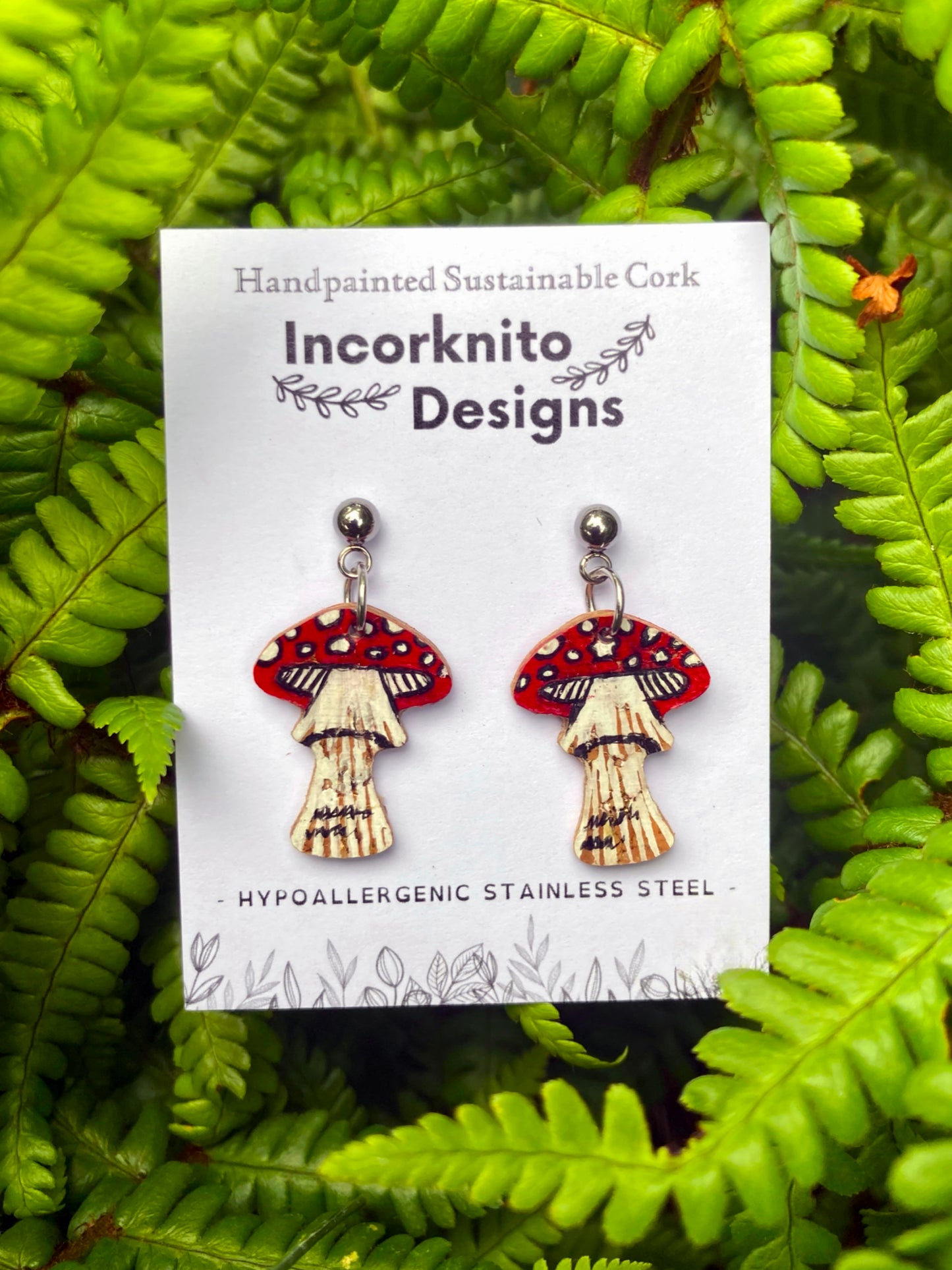 Fly agaric toadstool cork earrings on silver ball studs on Incorknito Designs card against lush green ferns — sustainable handpainted woodland earrings