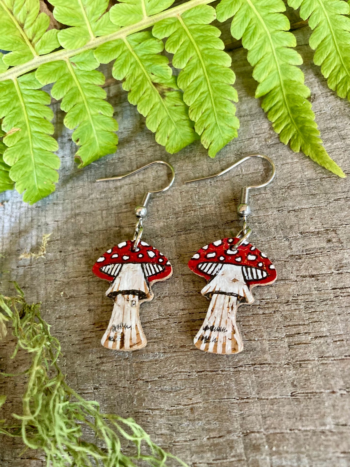 Handpainted fly agaric toadstool cork earrings on silver hooks flat lay on weathered wood with green fern and moss — eco-friendly woodland jewellery