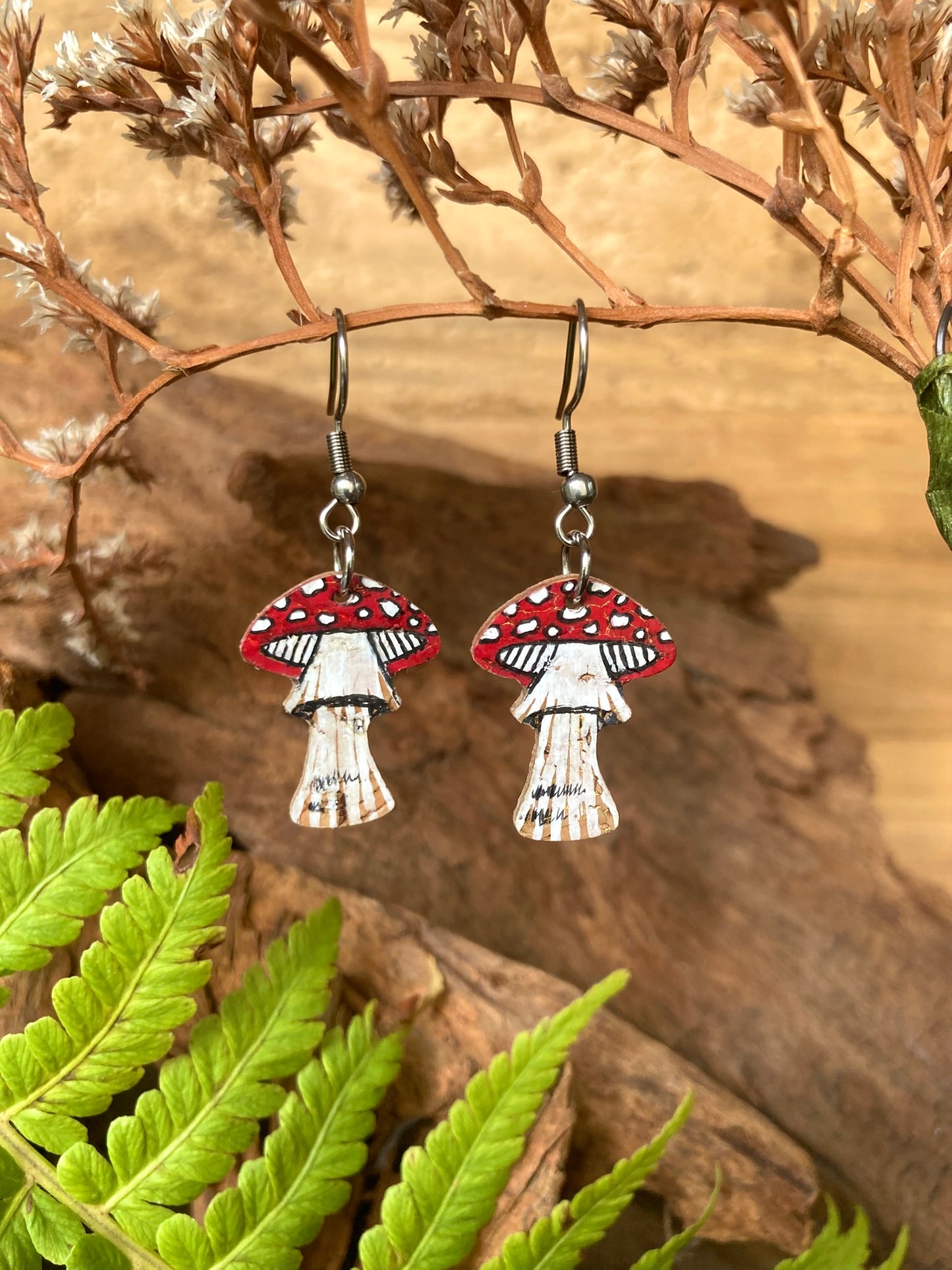 Fly agaric toadstool cork earrings on silver hooks hanging from dried russet botanicals with fern and driftwood — handpainted sustainable woodland jewellery