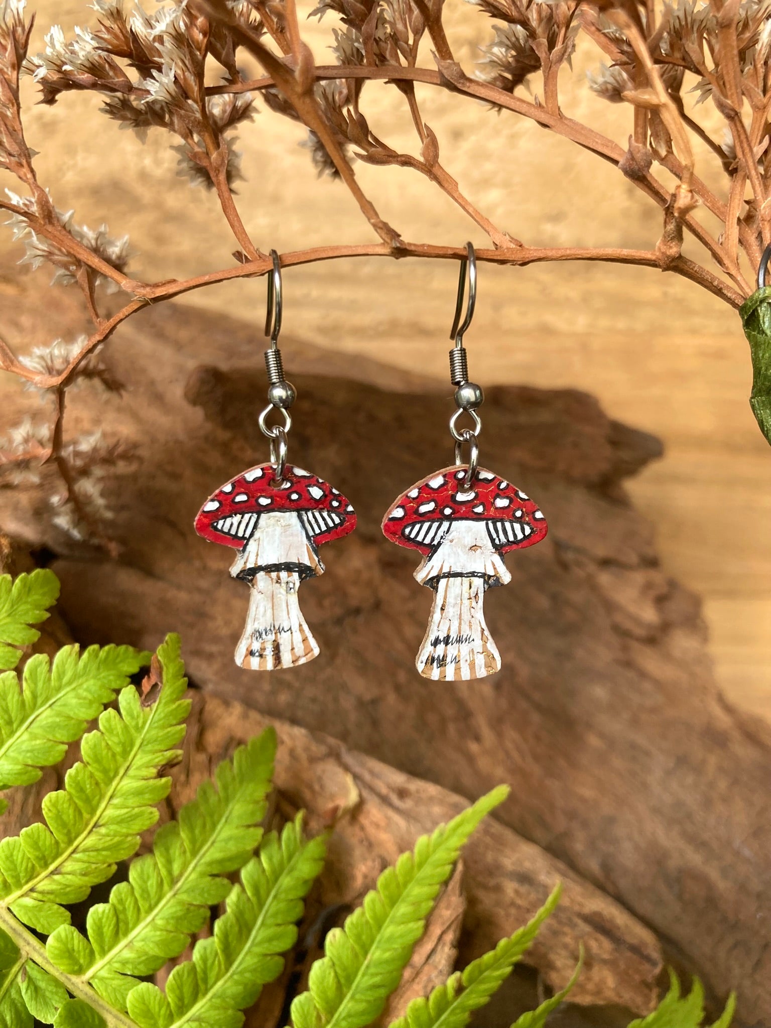 Fly agaric toadstool cork earrings on silver hooks hanging from dried russet botanicals with fern and driftwood — handpainted sustainable woodland jewellery