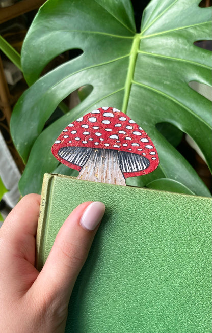 Fly agaric toadstool cork bookmark peeking out of a green hardback book held against a monstera leaf — handpainted sustainable cork