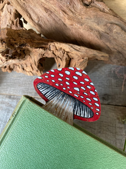 Fly agaric toadstool cork bookmark peeking out of a green hardback book with driftwood backdrop — showing double-sided design