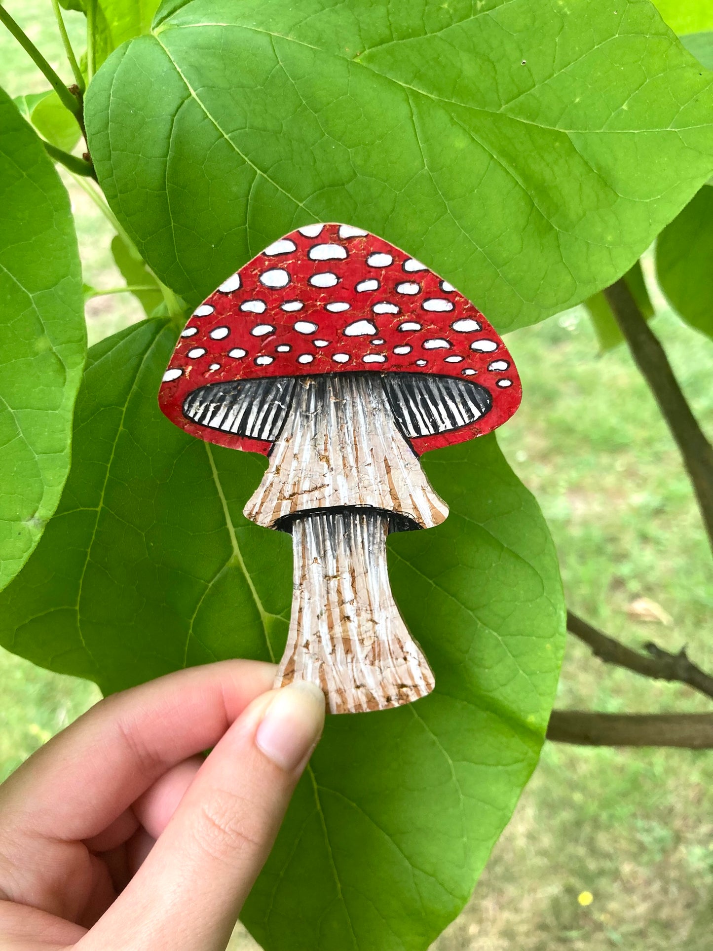 Handpainted fly agaric toadstool cork bookmark held against large green leaves — showing full bookmark with red cap, white spots and natural cork stem