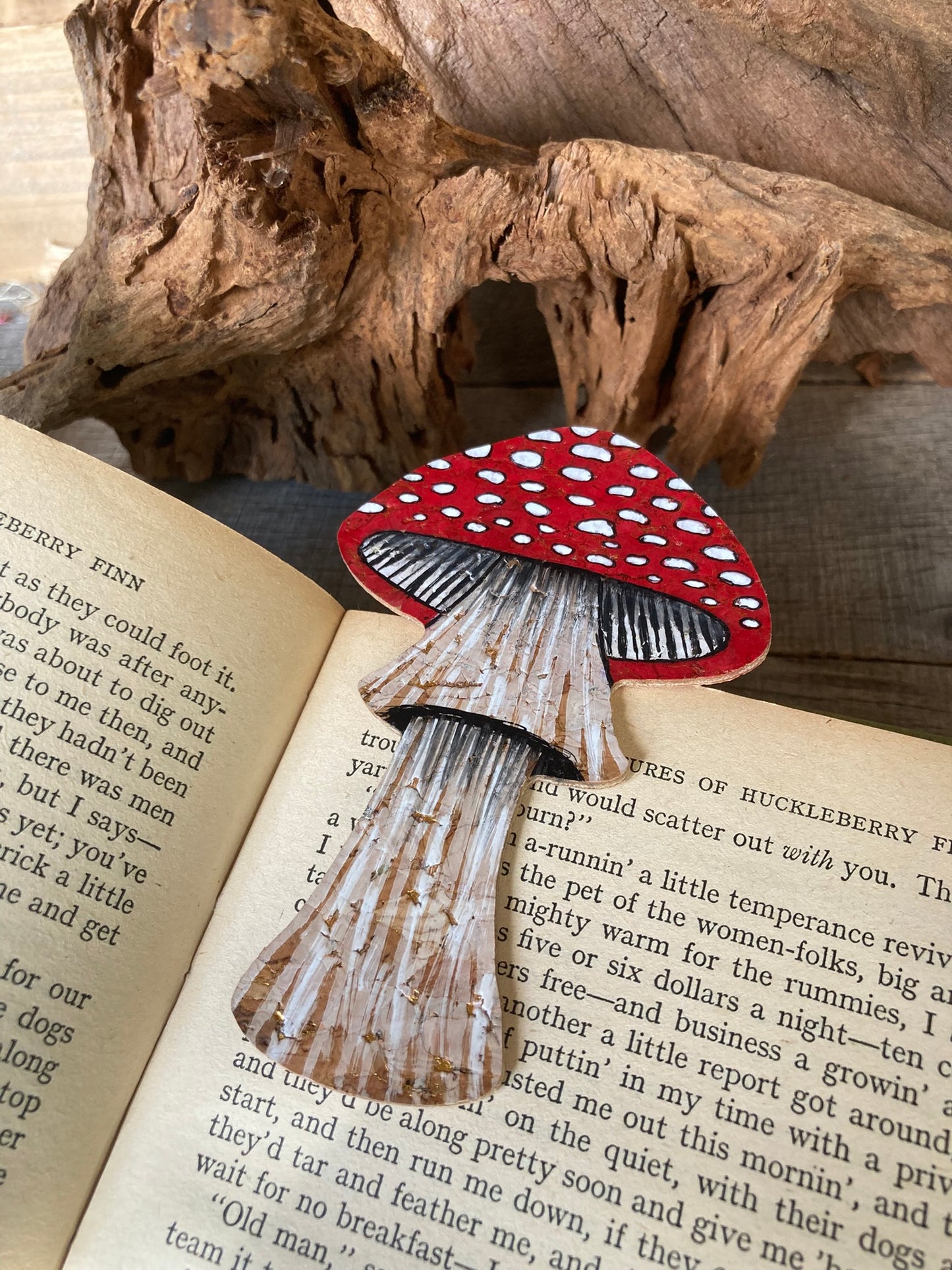 Fly agaric toadstool cork bookmark laid on open vintage book pages with driftwood backdrop — handpainted sustainable cork bookmark