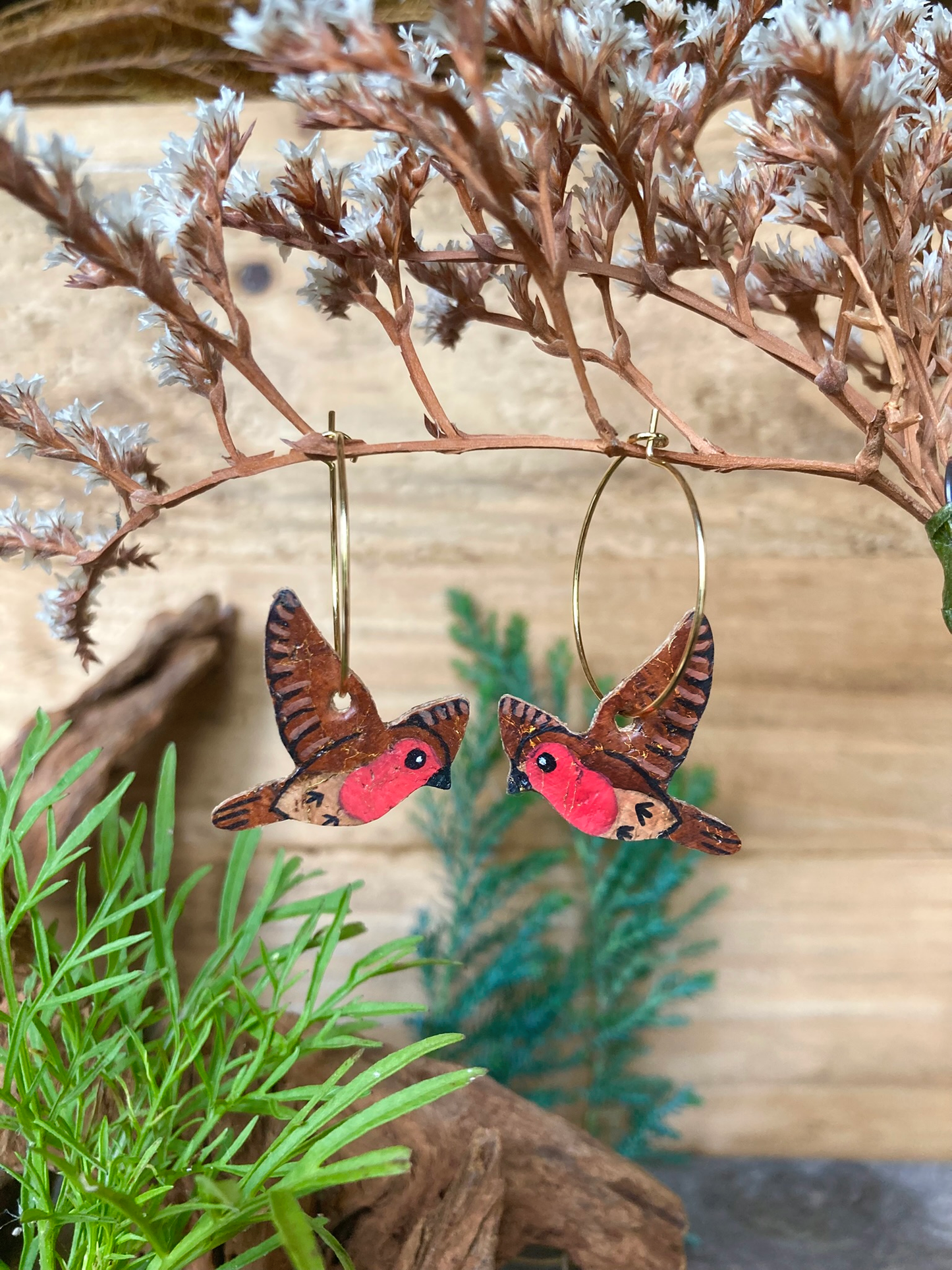 Handpainted flying robin cork earrings on gold hoops hanging from a dried floral branch — eco-friendly jewellery handmade in Suffolk