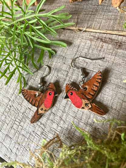 Flying robin cork earrings on silver hooks laid flat on weathered driftwood surrounded by moss and foliage
