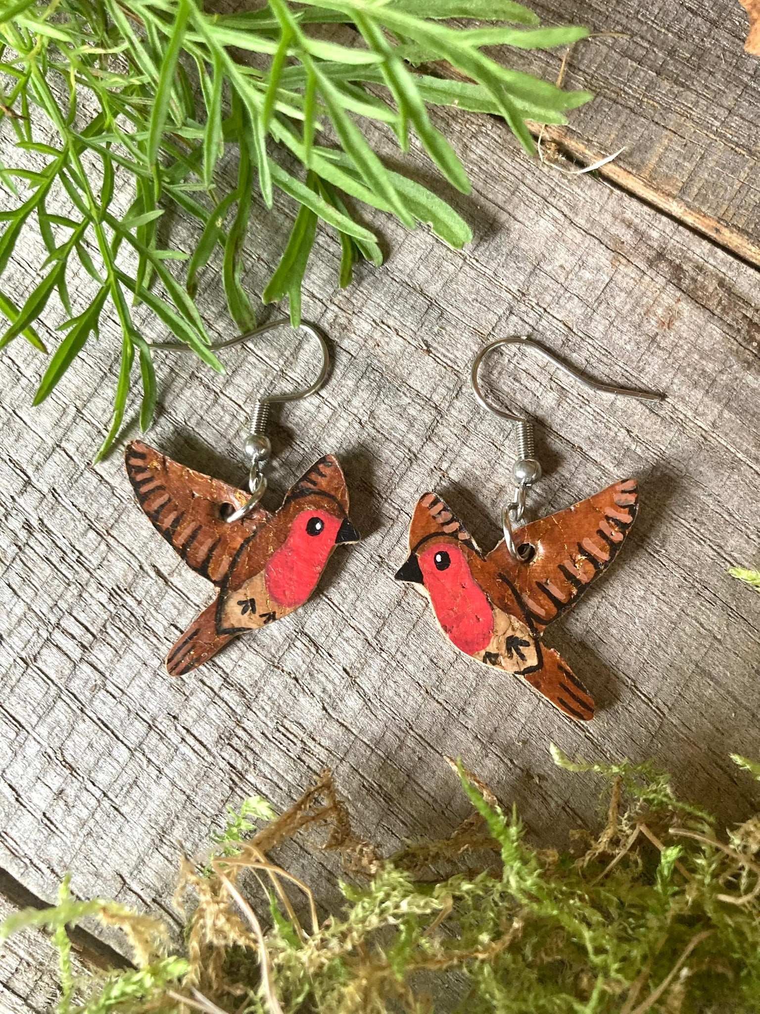 Handpainted flying robin cork earrings on silver hooks, flat lay on driftwood with moss and green foliage