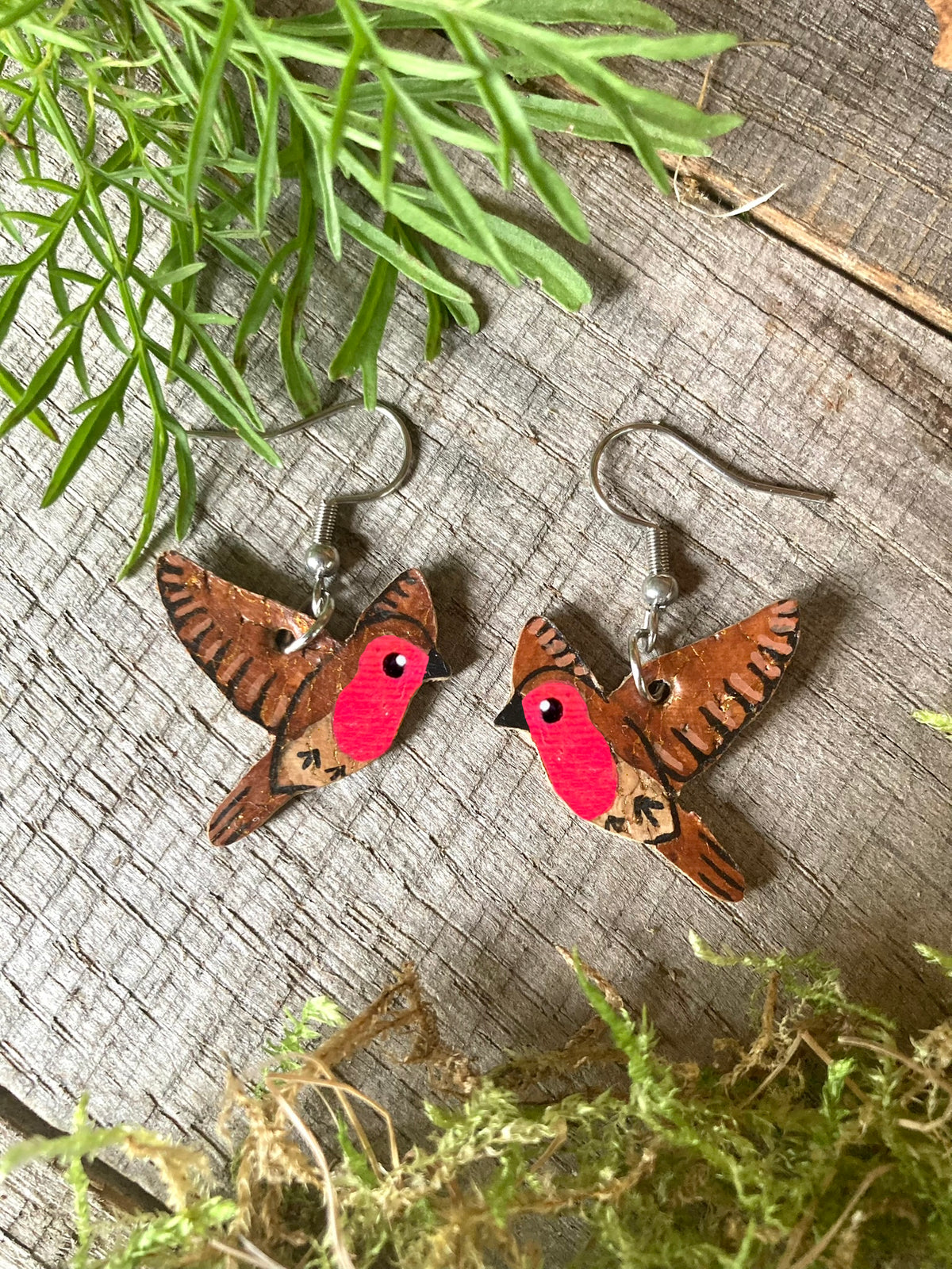 Handpainted flying robin cork earrings on silver hooks, flat lay on driftwood with moss and green foliage
