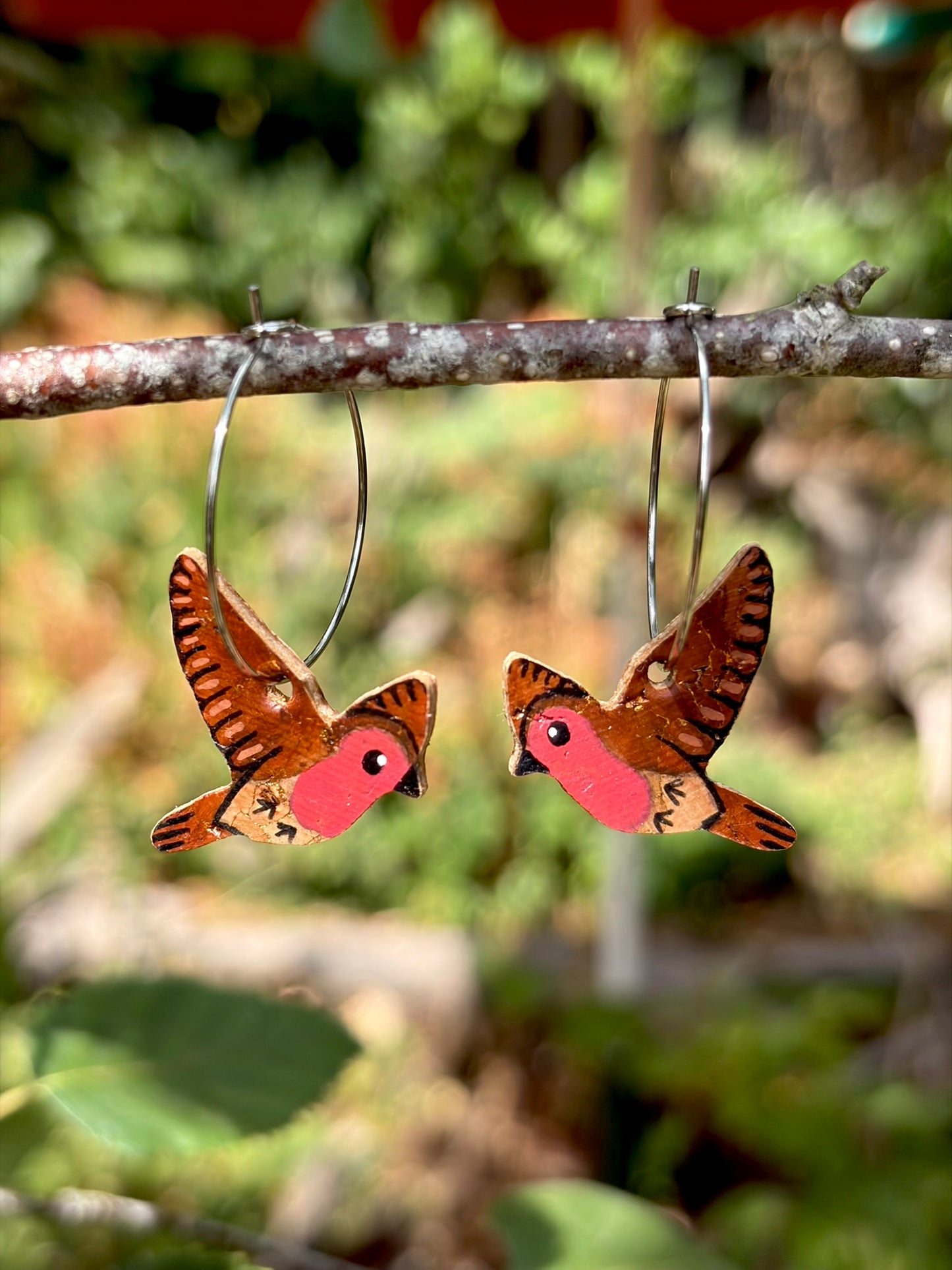 Handpainted flying robin cork hoop earrings hanging from a branch with blurred garden background