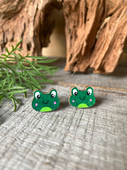 Handpainted frog cork stud earrings on a driftwood surface with wild greenery