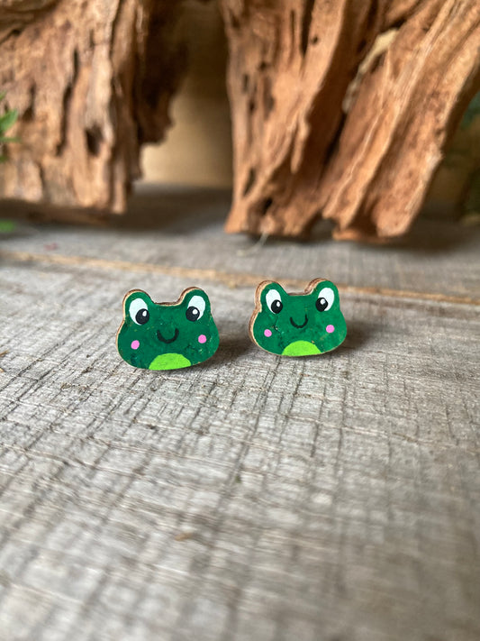 Handpainted frog cork stud earrings with green face, pink blushing cheeks and expressive eyes on a driftwood surface