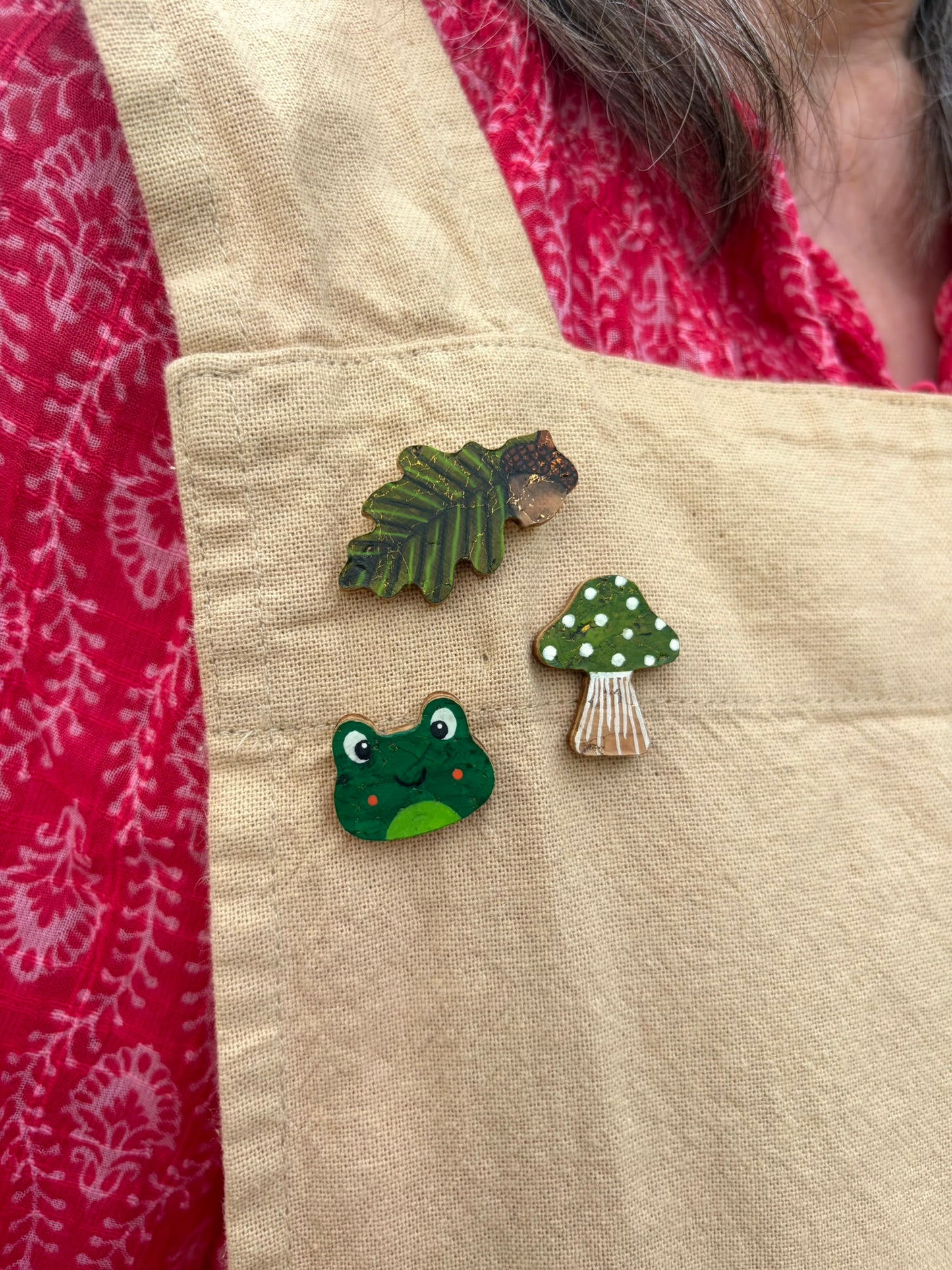 Frog, oak leaf, and toadstool cork pin badges pinned to a linen apron— handpainted sustainable badges from Incorknito Designs