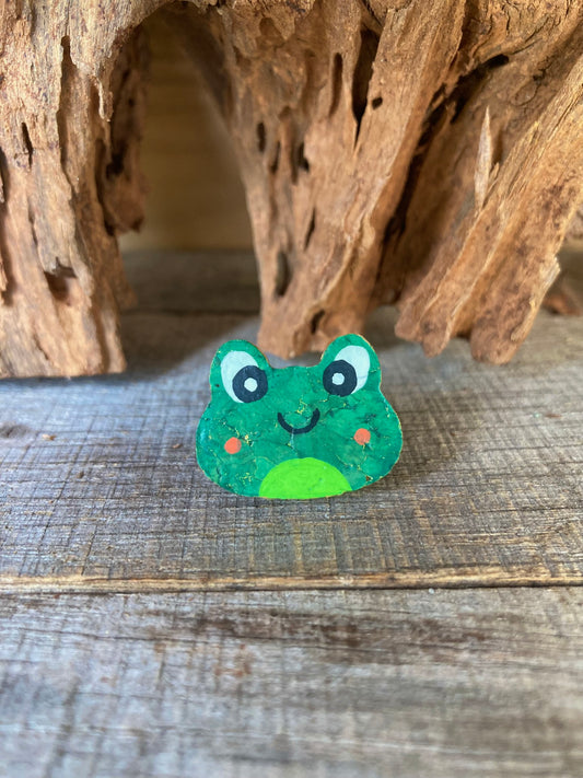 Handpainted smiley frog cork pin badge propped against driftwood — bright green frog face with rosy cheeks, handmade in Suffolk UK