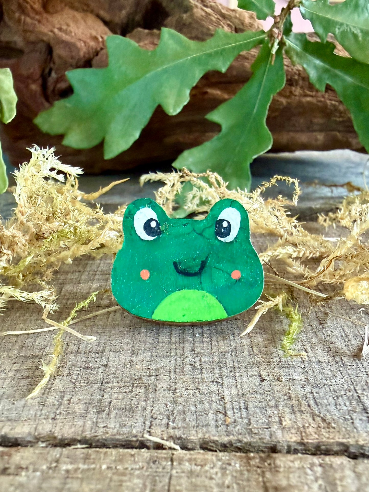 Handpainted orange cheeked frog cork pin badge face-on on weathered wood with dried moss and oak leaves — bright green frog with rosy orange cheeks