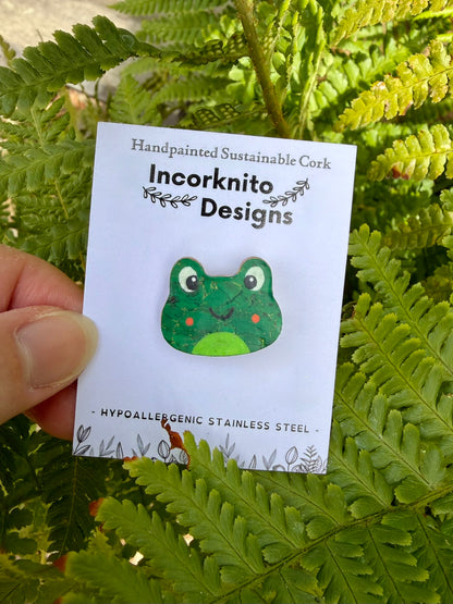 Orange cheeked frog cork pin badge on Incorknito Designs pin badge card held against green ferns