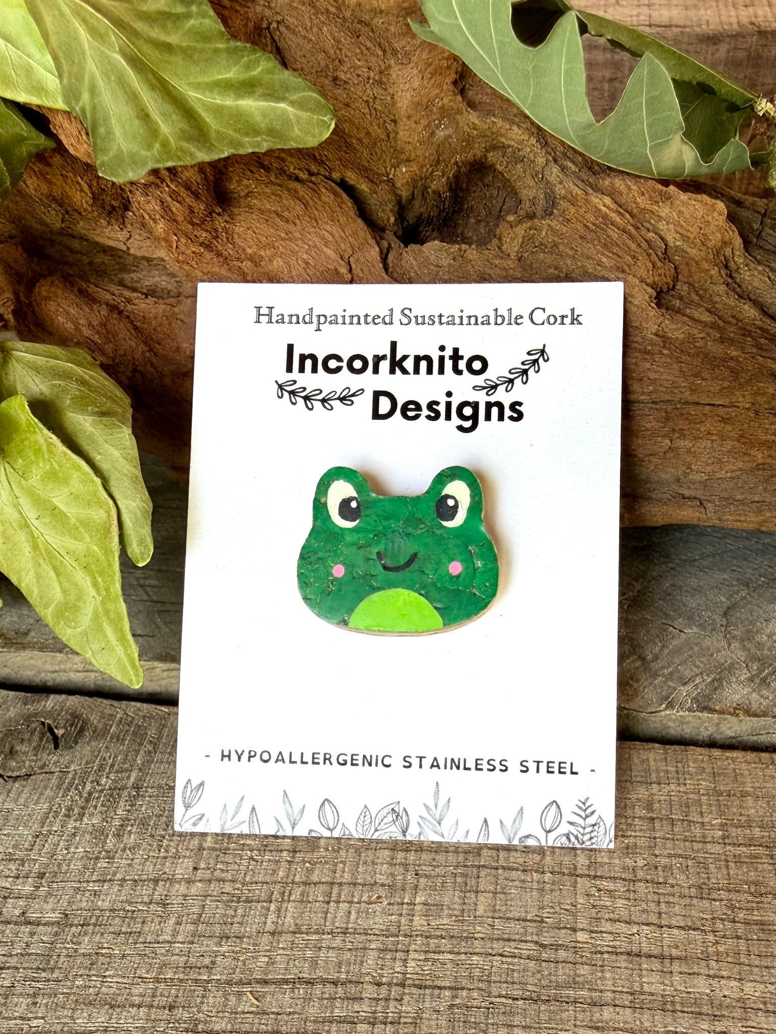 Pink cheeked frog cork pin badge on Incorknito Designs branded card against driftwood and oak leaves